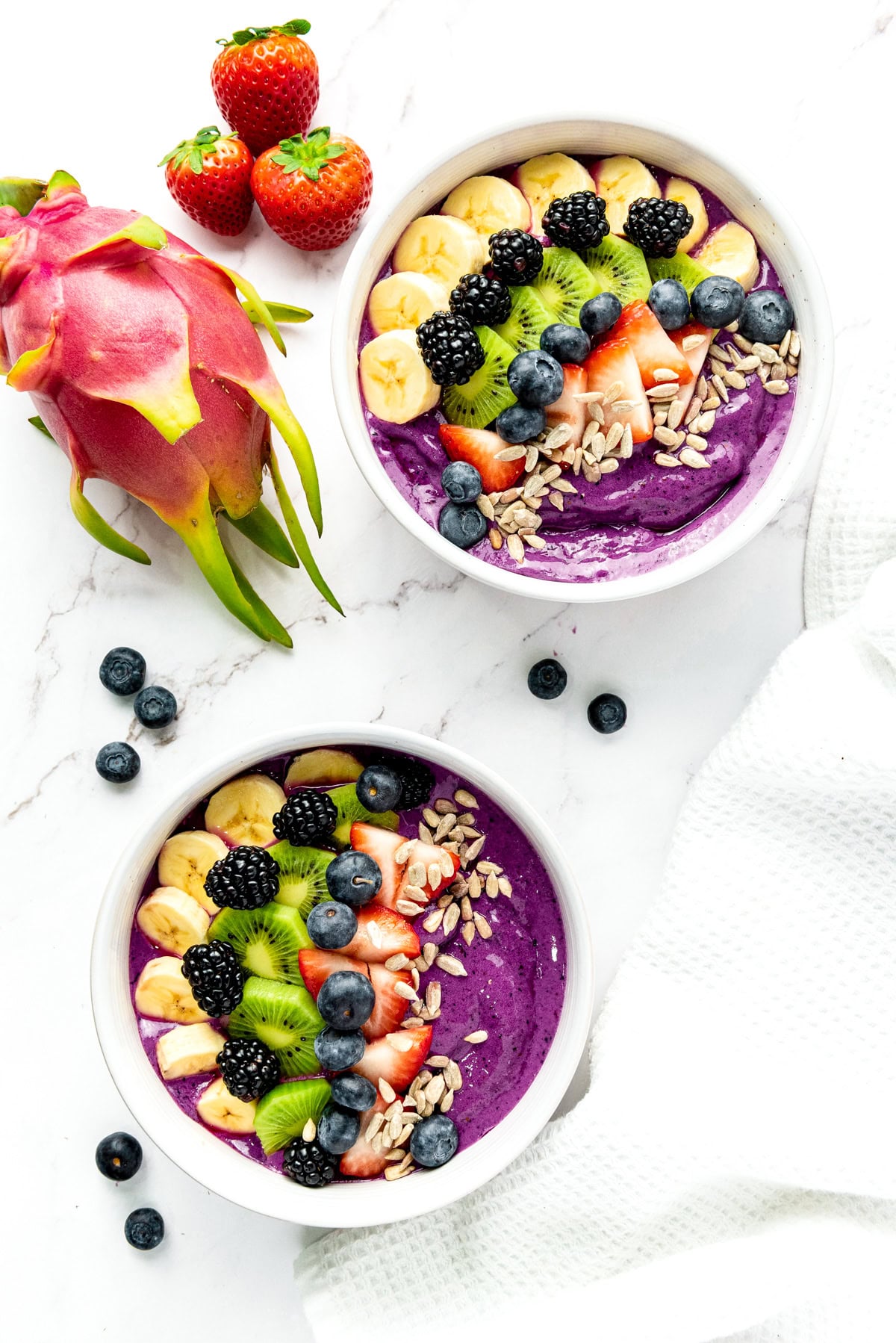 Two acai bowls topped with banana, kiwi, berries, and seeds, with strawberries and dragon fruit nearby.