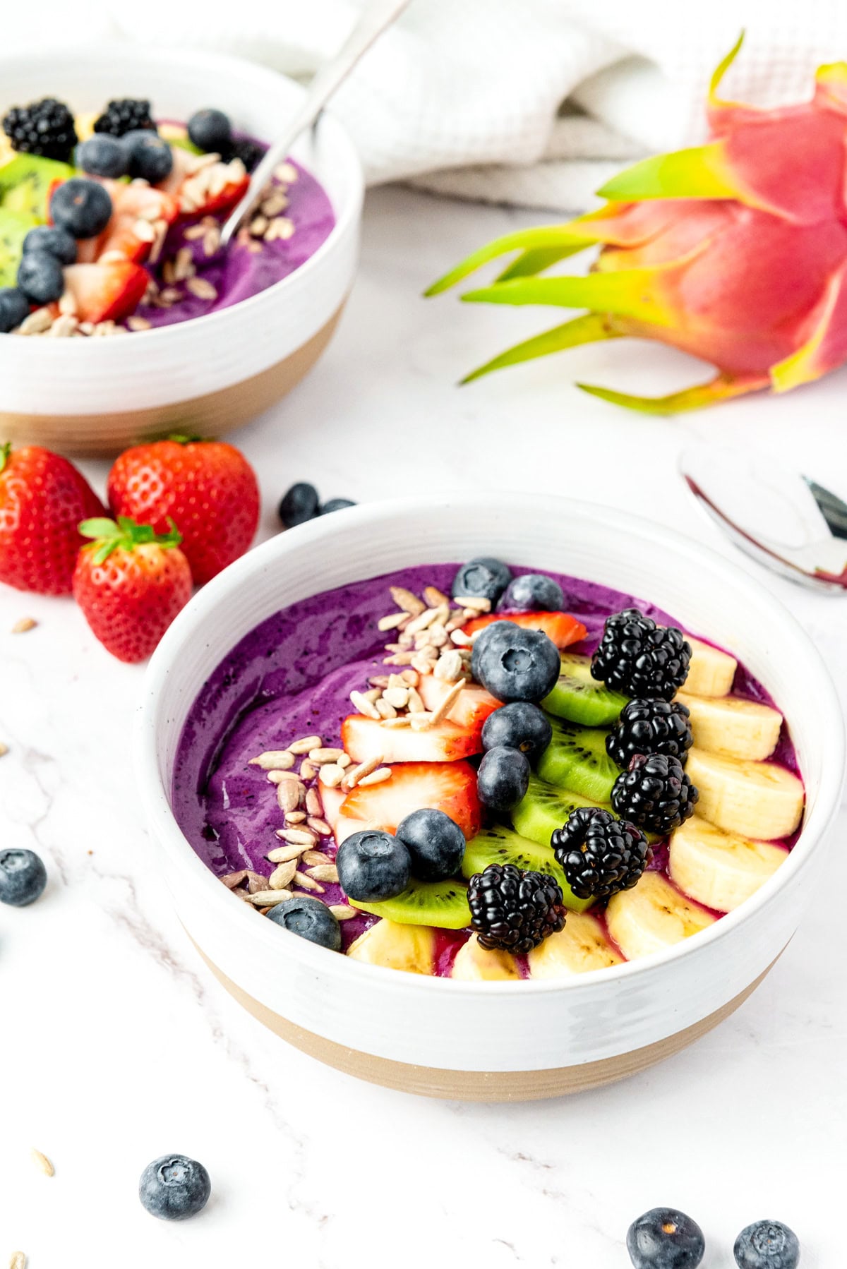 A smoothie bowl topped with banana, kiwi, berries, and seeds, with strawberries and dragon fruit nearby.