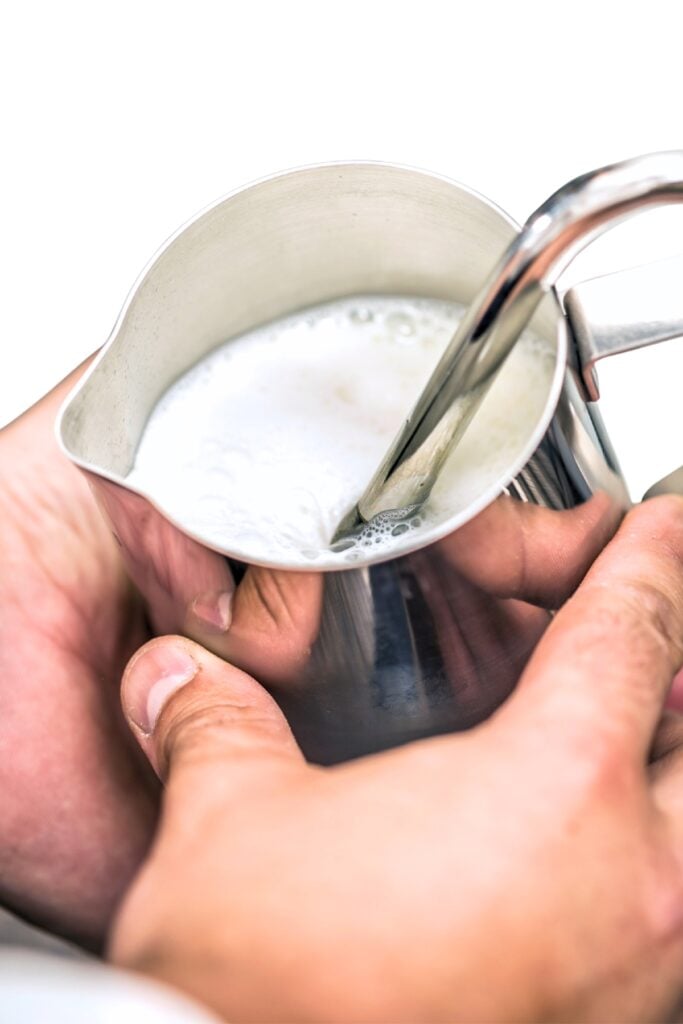 Close-up of hands frothing milk with a steam wand in a metal pitcher, white background.