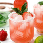 Pink cocktail with ice, sugar rim, basil, and strawberry garnish; lime and strawberries nearby.