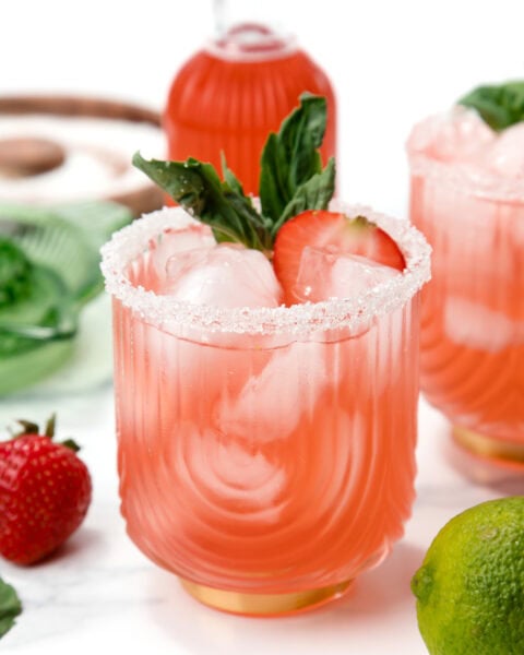 Pink cocktail with ice, sugar rim, basil, and strawberry garnish; lime and strawberries nearby.