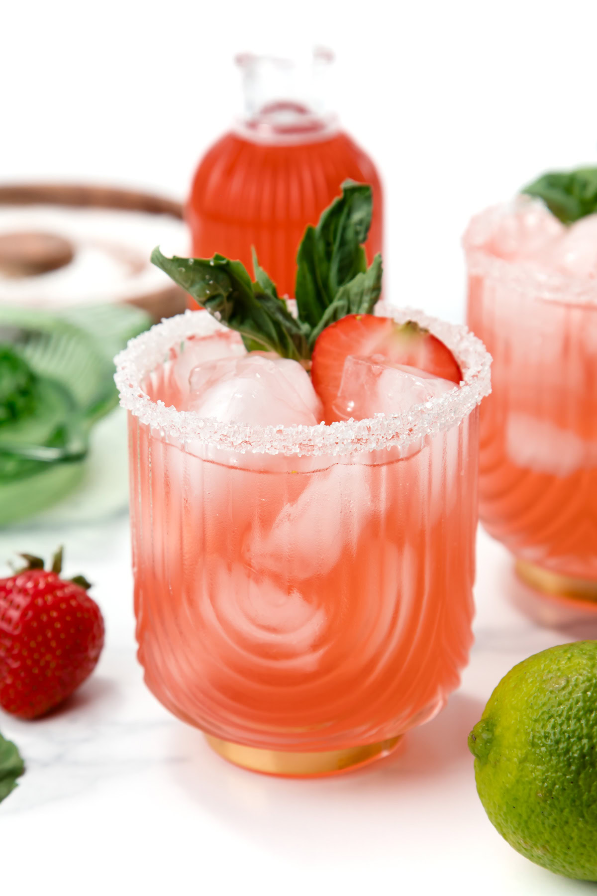 Pink cocktail with ice, sugar rim, basil, and strawberry garnish; lime and strawberries nearby.