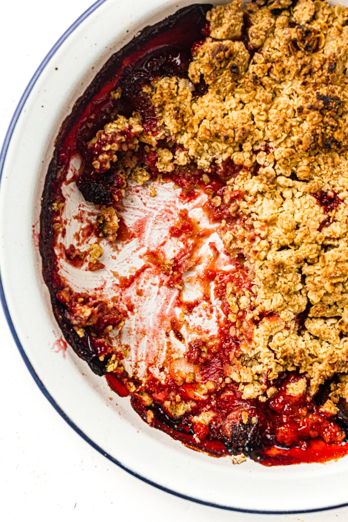 Partially eaten berry crumble dessert in a white dish, showing golden topping and red fruit filling.