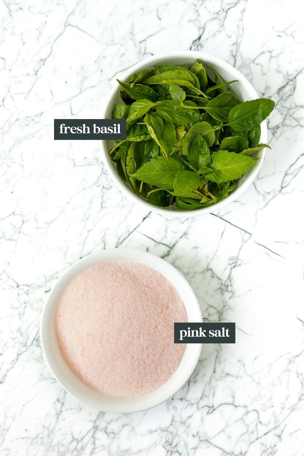 Bowl of fresh basil and bowl of pink salt on a white marble surface, each labeled.