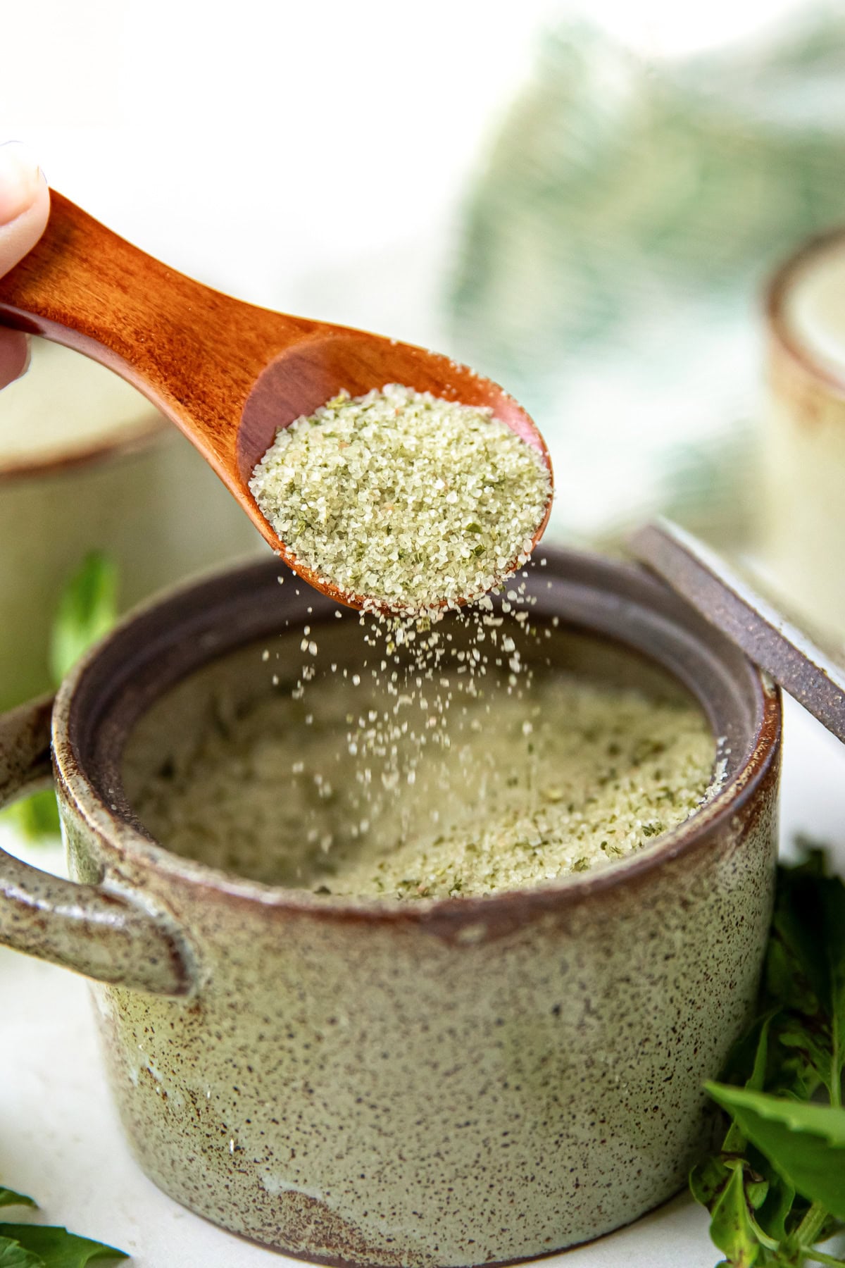 A wooden spoon sprinkles green herb salt into a ceramic bowl, with fresh herbs nearby.