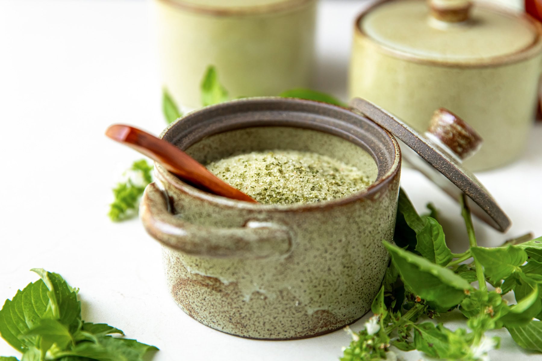 A ceramic jar with green seasoning inside and a wooden spoon, surrounded by fresh green herbs.