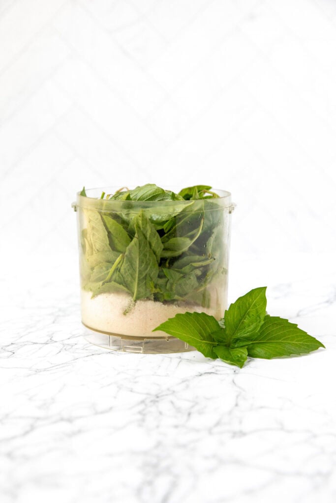 Fresh basil leaves and a light powder in a plastic container, with basil sprigs on a white marble surface.