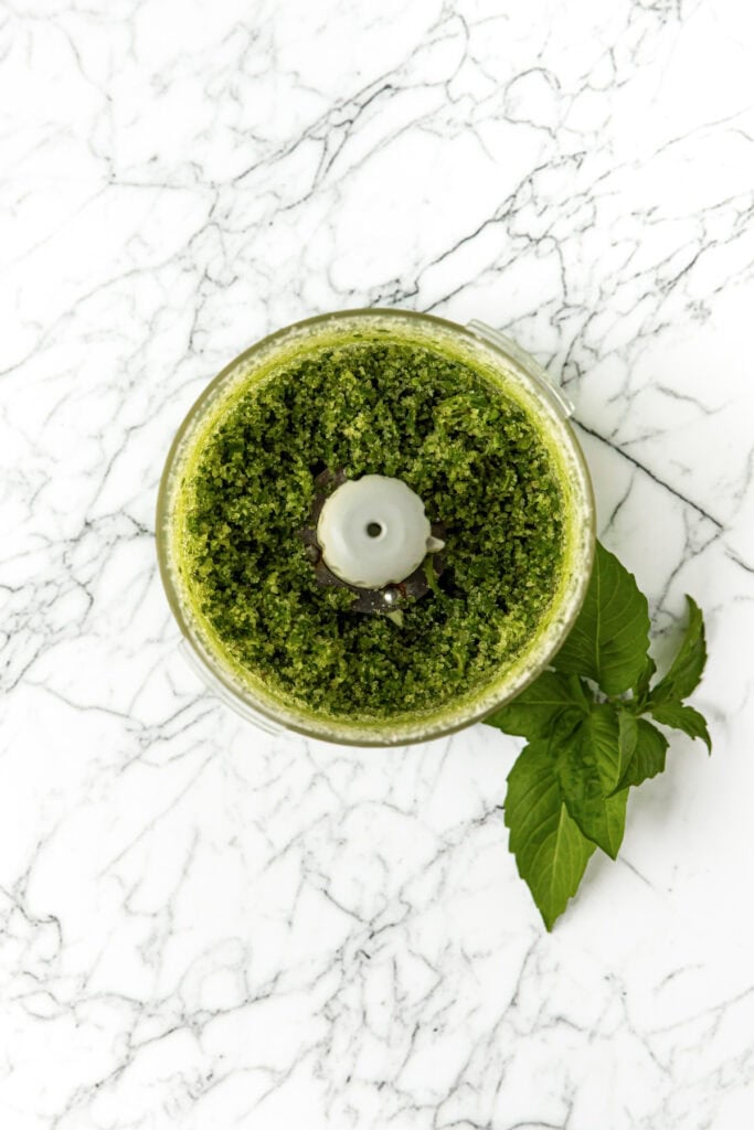 Top view of green pesto in a food processor on a marble surface with fresh basil leaves beside it.