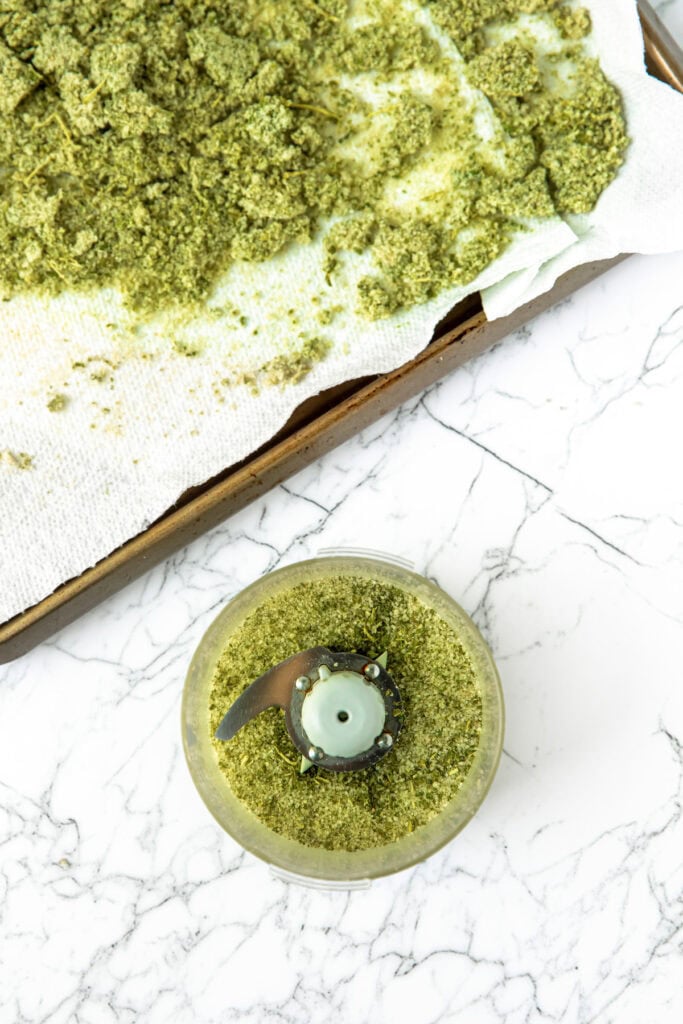 Tray with ground green herbs drying on paper towel and a food processor with herbs on a marble countertop.