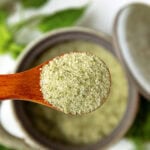 A wooden spoon holds green herb salt above a ceramic bowl, with fresh herbs in the background.