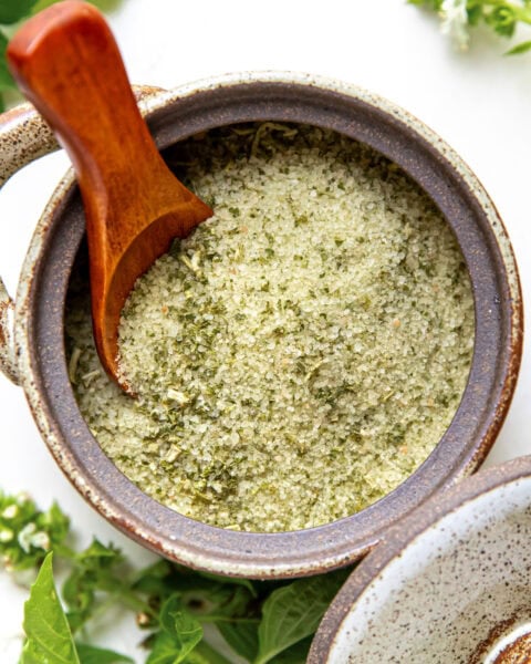 Ceramic jar filled with green herb salt, wooden spoon inside, fresh basil leaves around.