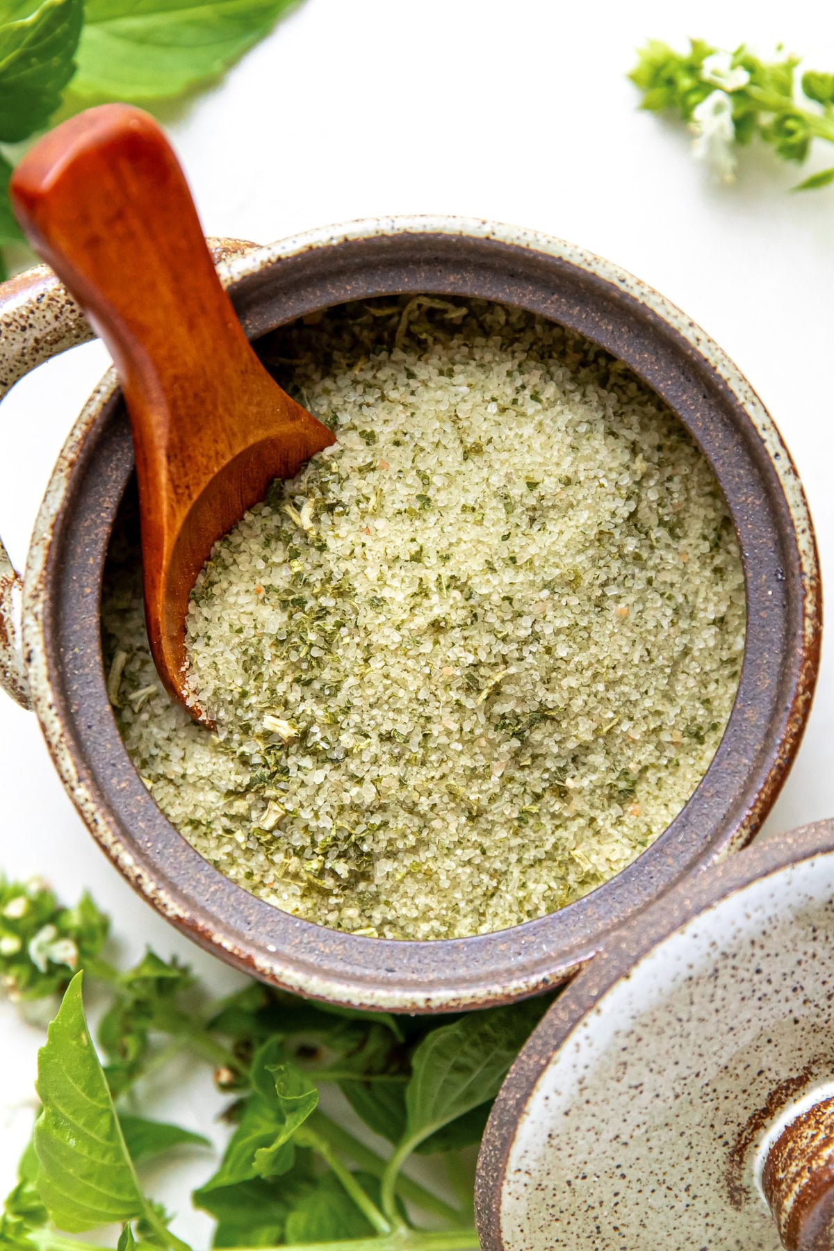 Ceramic jar filled with green herb salt, wooden spoon inside, fresh basil leaves around.