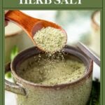 A wooden spoon sprinkles homemade herb salt into a rustic ceramic bowl, with herbs in the background.