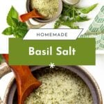 Top-down view of a pottery jar filled with homemade basil salt, a wooden spoon, and fresh basil leaves.