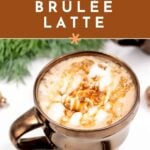 Caramel brûlée latte in a brown mug with whipped cream and caramel drizzle, holiday decor in background.