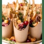 Individual paper cups filled with cheese, meats, pretzels, grapes, and herbs, arranged on a tray.