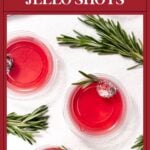 Three red jello shots in cups, garnished with rosemary and cranberries, on a white surface.