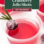 Plastic cup of red cranberry jello with sugared cranberry and rosemary garnish, labeled as cranberry jello shots.