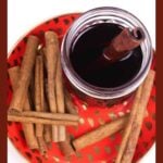 A jar of dark whiskey with a cinnamon stick, surrounded by cinnamon sticks on a red tray.