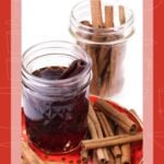 Two jars, one with cinnamon sticks and one with dark whiskey, sit on a plate with more cinnamon sticks.