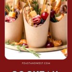 Brown paper cups filled with assorted cocktail appetizers, cheese, breadsticks, and herbs on a white plate.