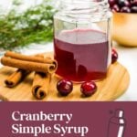A jar of cranberry syrup with cinnamon sticks, cranberries, and pine on a wooden board; holiday drink ad.