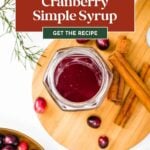 Jar of cranberry syrup on cutting board with cinnamon sticks, cranberries, and pine sprigs around it.