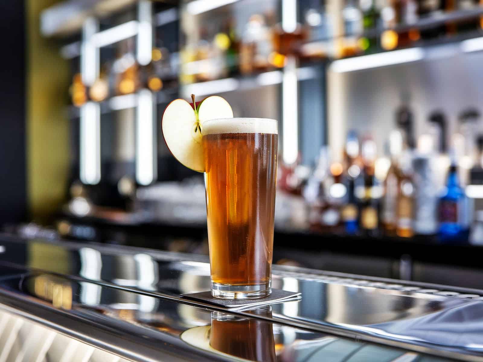 A glass of cider with a frothy head and apple garnish sits on a bar counter with bottles in the background.