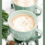 Two green mugs filled with eggnog latte, topped with whipped cream, next to holiday ornaments and greenery.