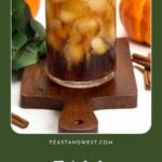 Iced coffee in a glass on a wooden board, with pumpkins, leaves, and cinnamon sticks in the background.