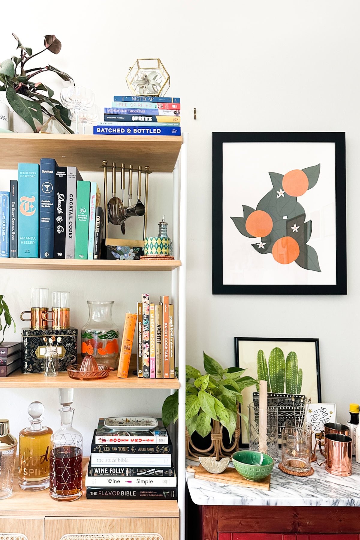 Bookshelf and bar cart with plants, books, glassware, and framed orange fruit art on a white wall—perfect for those looking to stock your bar with the best alcohol alongside curated décor.