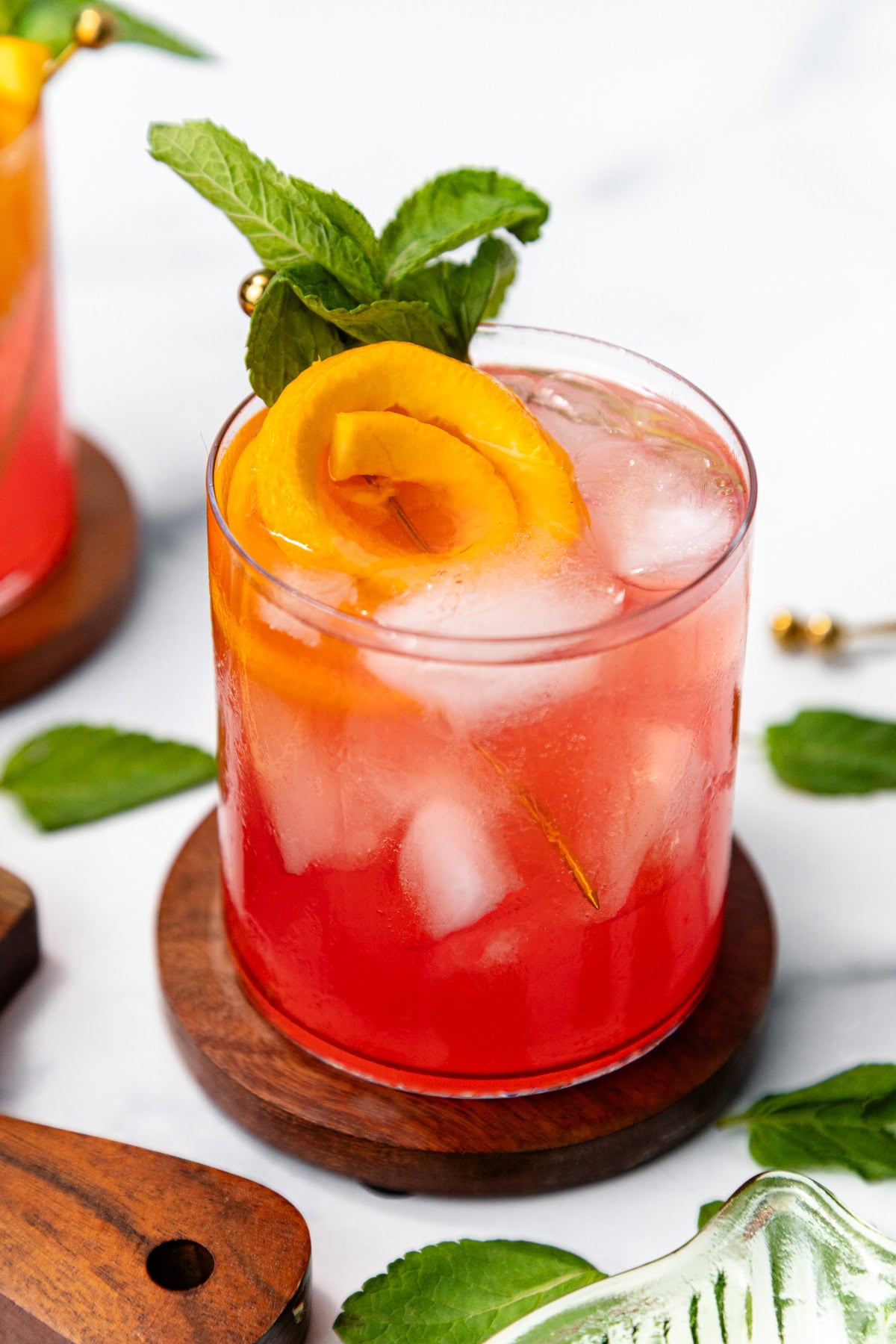 A red cocktail with ice, an orange twist, and mint garnish in a glass on a wooden coaster.