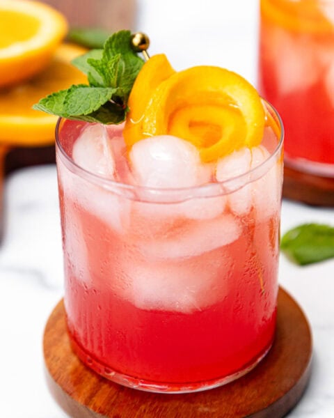 Pink cocktail with ice, orange peel garnish, and mint sprig in a glass on a wooden coaster.