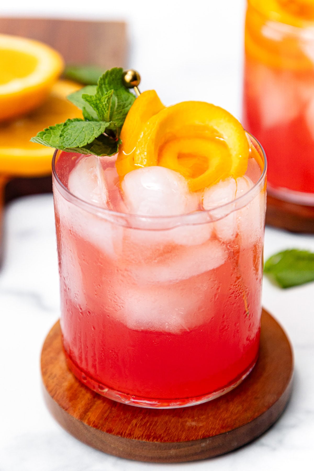 Pink cocktail with ice, orange peel garnish, and mint sprig in a glass on a wooden coaster.