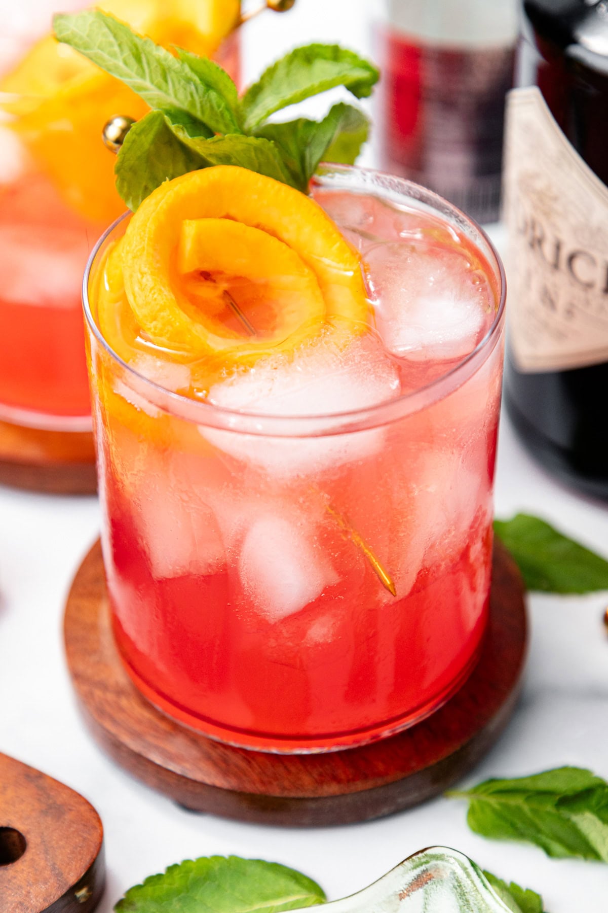 A red cocktail with ice, orange peel, and mint garnish in a glass on a wooden coaster.