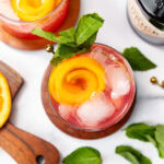 A cocktail with ice, orange peel, and mint garnish, seen from above. Mint leaves and orange nearby.