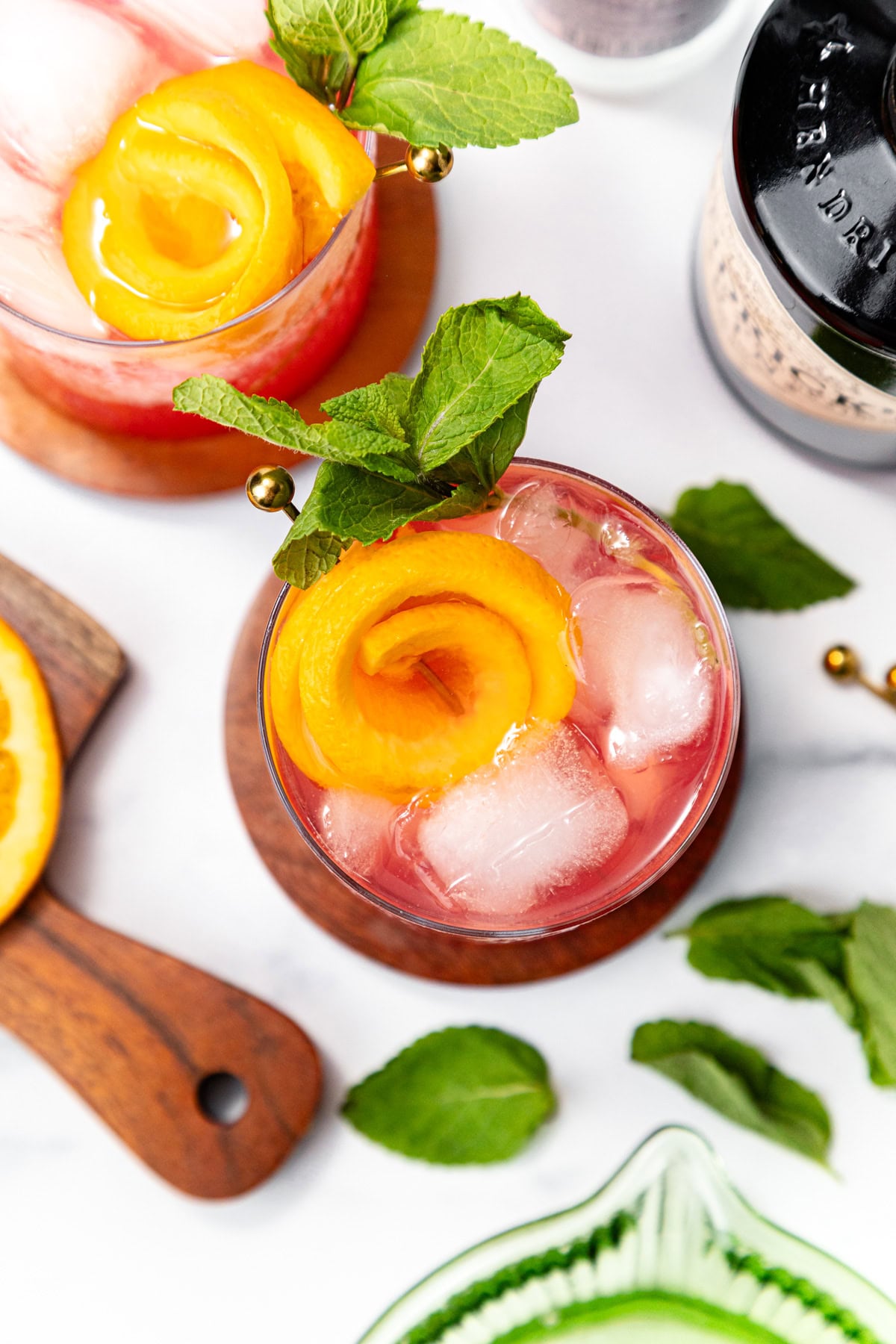 A cocktail with ice, orange peel, and mint garnish, seen from above. Mint leaves and orange nearby.