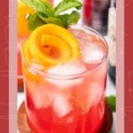 A pink gin daisy cocktail with ice, orange peel, and mint garnish on a wooden coaster.