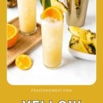 Two yellow cocktails with pineapple and orange garnish, beside a shaker and pineapple slices on a white table.