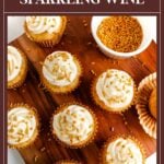 Seven cupcakes with white frosting and gold sprinkles on a wooden board near a bowl of sprinkles.