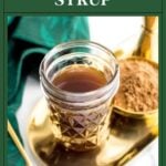 A jar of Irish cream syrup on a gold tray with cocoa powder, a spoon, and a green cloth.