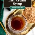 A glass of Irish cream syrup on a gold tray with cocoa powder, green cloth, and gold spoon nearby.