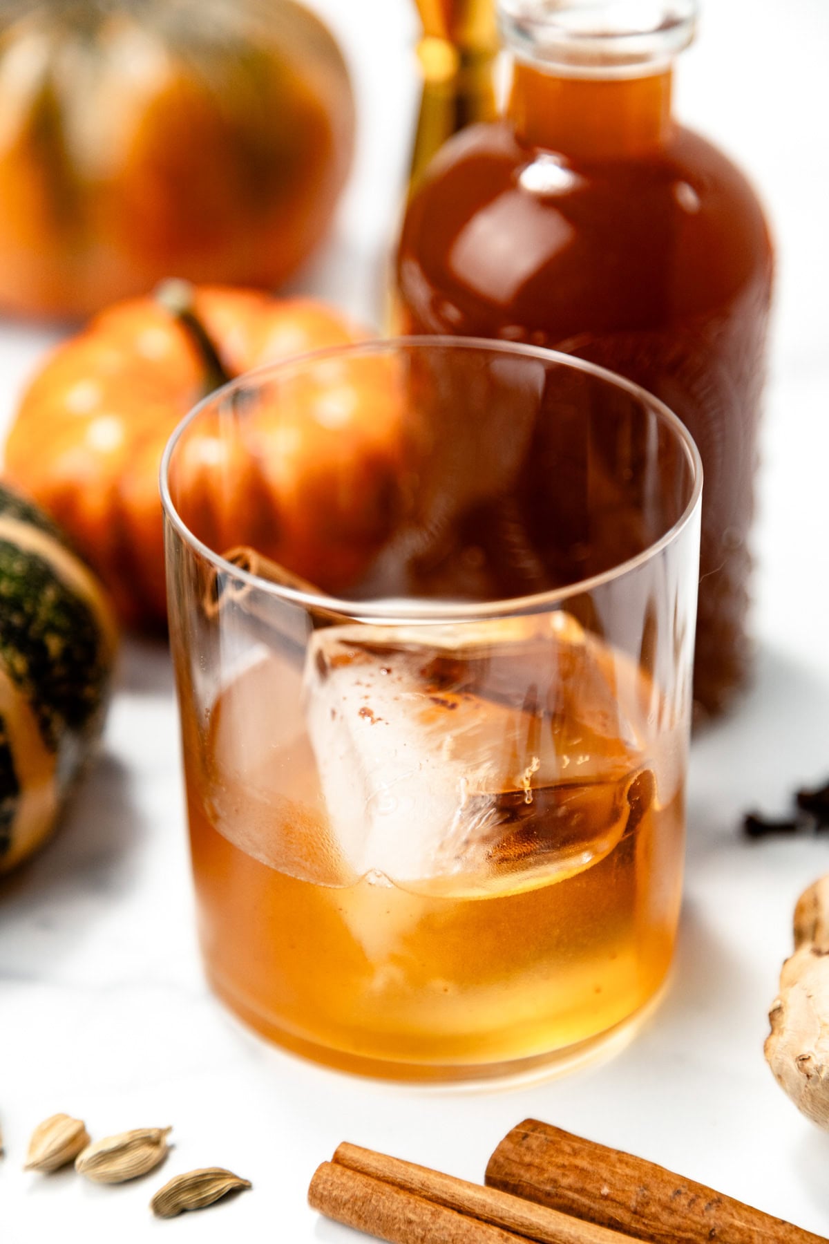 A glass of amber-colored drink with ice, surrounded by pumpkins, spices, and a bottle in the background.