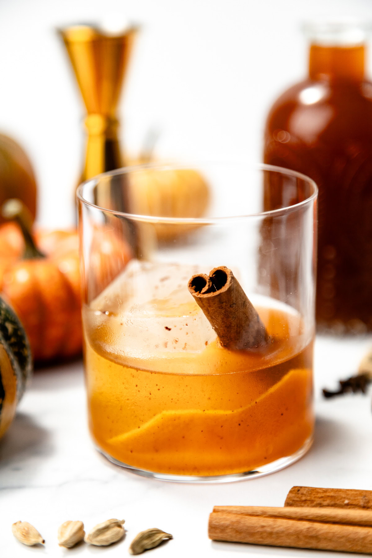 A glass of orange cocktail with a cinnamon stick, surrounded by pumpkins and spices on a white surface.