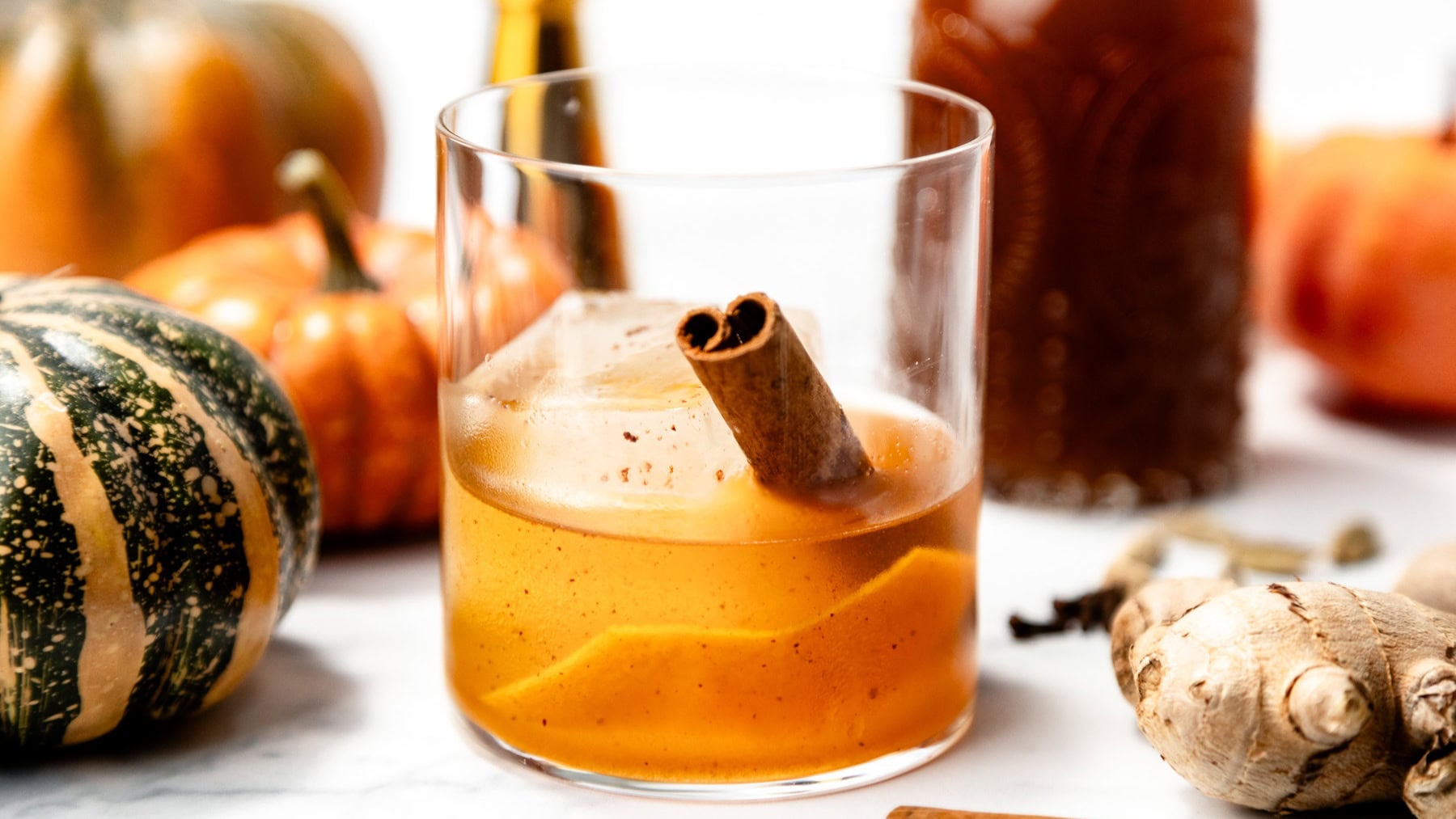 A glass of orange cocktail with ice and a cinnamon stick, surrounded by pumpkins and ginger root.
