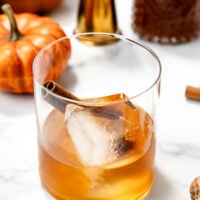 A glass of amber drink with ice and a cinnamon stick, surrounded by pumpkins and bottles on a white surface.