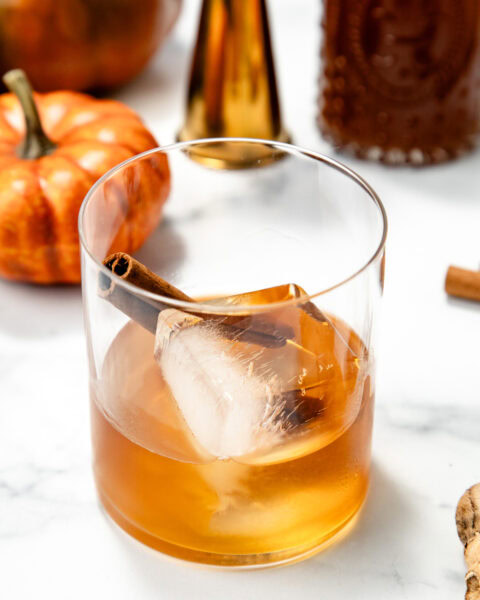 A glass of amber drink with ice and a cinnamon stick, surrounded by pumpkins and bottles on a white surface.