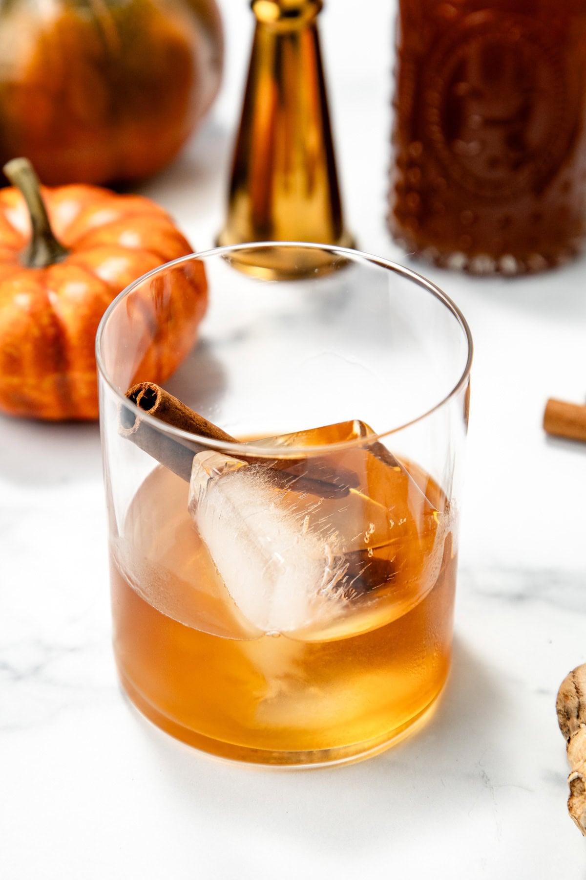 A glass of amber drink with ice and a cinnamon stick, surrounded by pumpkins and bottles on a white surface.