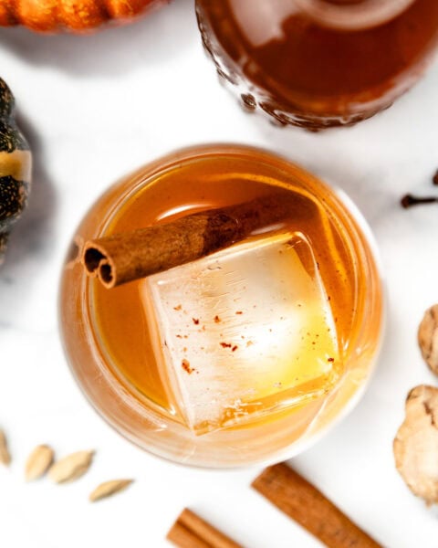 A glass of brown cocktail with a large ice cube and cinnamon stick, surrounded by spices and a pumpkin.
