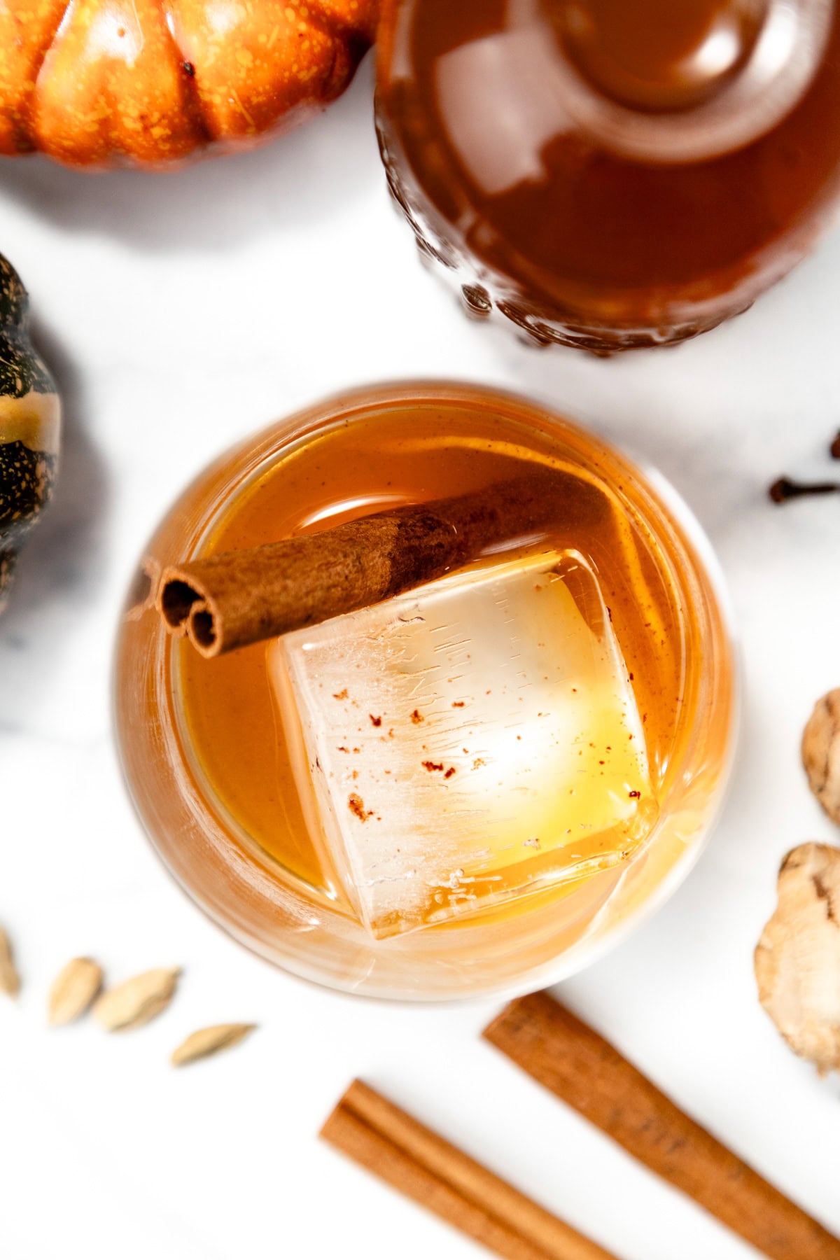 A glass of brown cocktail with a large ice cube and cinnamon stick, surrounded by spices and a pumpkin.
